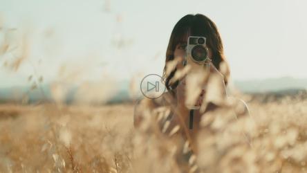 Behind wheat grass a woman films with a vintage 8mm camera in nature[]