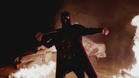 Angry vandal stands in front of a burning car during civil unrest[]
