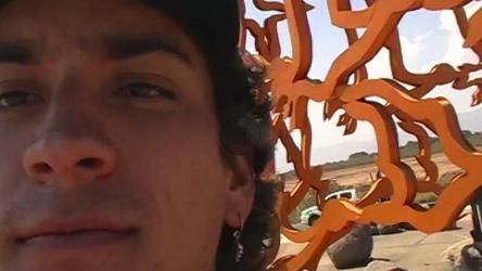 一个戴着卡车司机帽的年轻人正在拍摄与女友的接吻，背景是一个艺术的红色结构和草地。[免费商用]