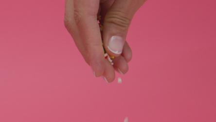 一个女人在甜甜圈上撒彩色的糖霜快餐 Fast Food:一个女人的特写镜头'；在粉红色的背景下，她的手在盘子里的一个上釉的甜甜圈上撒上了五颜六色的小水滴。