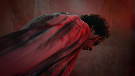 虐待狂折磨者触摸受害者的脸血腥 Gore:穿着考究的折磨者走到几乎昏迷的受害者面前，伸手抬起他的头。凶手身后黑暗而令人不安的房间里，一道昏暗的光线照耀着他。