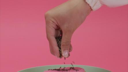 在甜甜圈上撒巧克力片快餐 Fast Food:一个女人'；s将巧克力片放在粉红色糖霜的甜甜圈上，放在粉红色背景的盘子里。