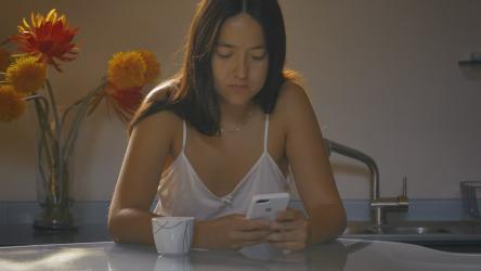 早上喝咖啡时用手机的女人饮食 Food:早上，一位女士在厨房的餐厅里，一边用杯子喝咖啡，一边聚精会神地查看手机，身后有鲜花和窗户。