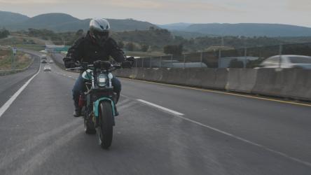 一个骑自行车的人在高速公路上加速摩托车 Motorcycle:一个穿着黑色夹克、蓝色牛仔裤和白色头盔的人在高速公路上骑着摩托车，汽车从道路两侧驶过。背景是绿色的山丘和晴朗的蓝天。