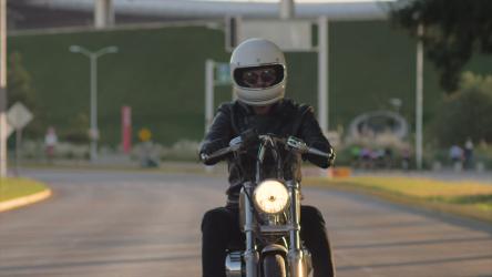 在孤独的街道上驾驶摩托车的人摩托车 Motorcycle:一个穿着黑色皮夹克、戴着白色头盔的人开着一辆哈雷戴维森摩托车在一条空荡荡的路上行驶。背景中的草和树。